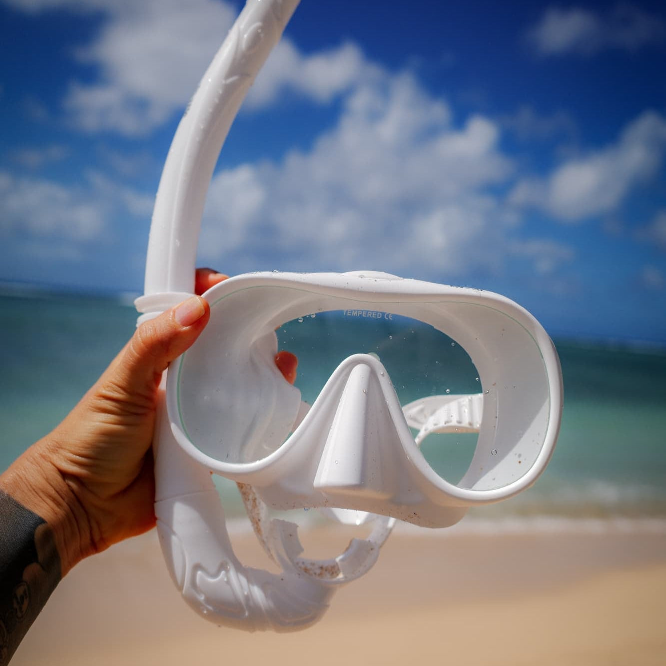 White snorkel mask held by a hand on a beach with blue sky and ocean in the background