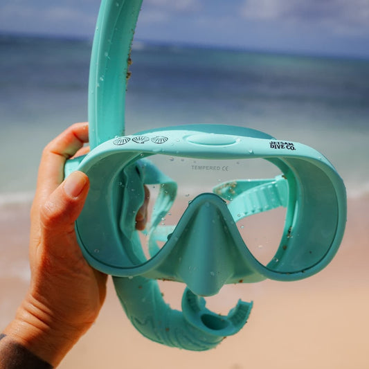 Turquoise snorkel mask held by a hand on a beach with ocean in the background