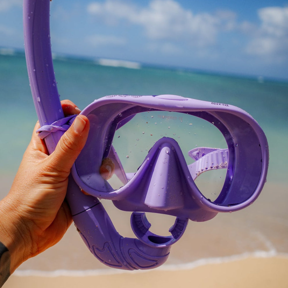 Purple diving mask held by a hand on a beach with ocean and sky in the background