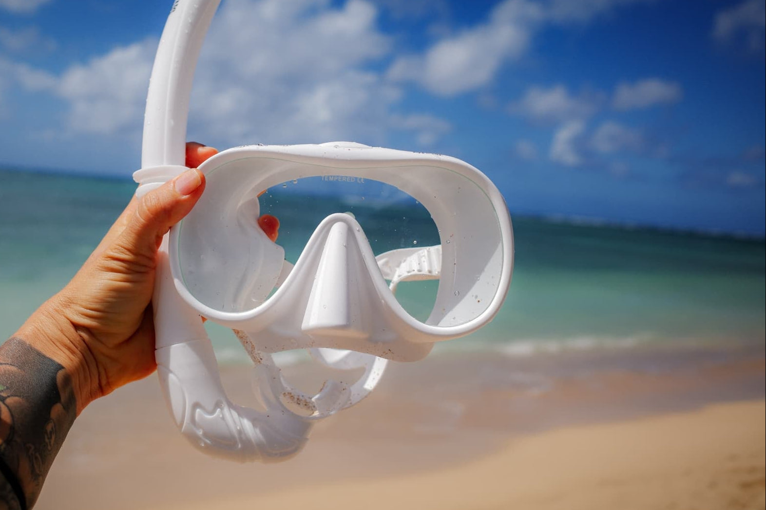 White diving mask held by a hand on a beach with ocean and sky in the background