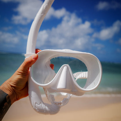 White snorkel mask held by a hand on a beach with blue sky and ocean in the background