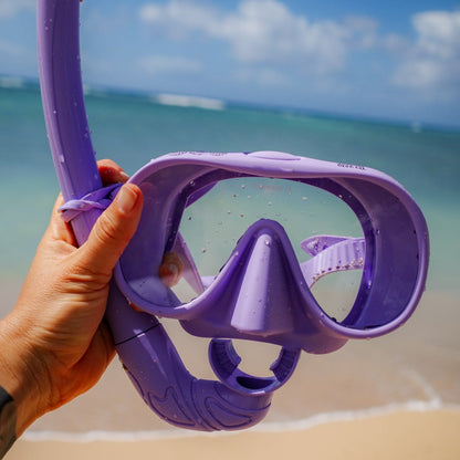 Purple diving mask held by a hand on a beach with ocean and sky in the background